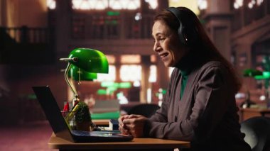 Yaşlı kadın üniversite öğrencisi Webinar 'da profesörle bilgisayar başında iletişim kuruyor, klasik akademik araştırmalarla modern telekomünikasyon kütüphanesinde okuyor. Seminer dersi. Kamera B.