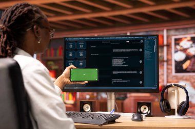 Ana ofisteki bilgisayar ekranında izole ekran akıllı telefonu ve AI LLM sohbet robotu kullanan bir kadın. Krom anahtar telefonu olan kişi monitörde yapay zeka sohbet robotu kullanıyor.
