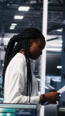 Yapay zeka sistemlerini yöneten dikey video veri merkezi mühendisleri, evrak dosyalarını inceliyorlar. Çok ırklı sunucu merkezinde evrak okuyan Afrikalı Amerikalı kadın ve Asyalı erkek