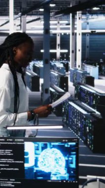 Yapay zeka sistemlerini yöneten dikey video veri merkezi mühendisleri, evrak dosyalarını inceliyorlar. Çok ırklı sunucu merkezinde evrak okuyan Afrikalı Amerikalı kadın ve Asyalı erkek