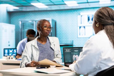 Siyahi pratisyen hekim ve hasta bir klinikte sağlık kontrolü ve tedavi seçeneklerini tartışıyor, iletişim ve tıbbi uzmanlığı vurguluyor. Kadın uzman tavsiye veriyor.