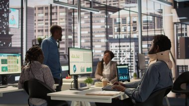 Siyahi işadamı, boyun kırığıyla, korunma ve destek için rahim boynu tasması takarken, acıyla başa çıkmak için masa başında çabalıyor. Yaralı işçi görevleri çözmeye çalışır. Kamera B.