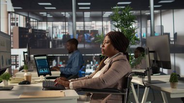 Finansal strateji üzerinde çalışan, organizasyonel amaçlara ulaşmak için veri analizlerini gözden geçiren Afrikalı Amerikalı bir çalışan. Kadın analist maluliyet dostu bir ofiste çalışıyor. Kamera B.