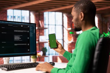 Evde elinde akıllı telefon tutan adam AI LLM sohbet robotu asistanına sorular soruyor. Yeşil ekranlı telefon kullanarak yapay zeka robotundan faydalı bilgiler alan kişi