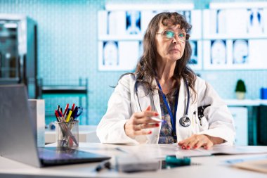 Deneyimli bir doktor, erkek bir hastayla görüşürken notlar alır, daha eski muayeneler ve test sonuçları için hastane kayıtlarını kontrol eder. Doktor muayenehanede ilaç tavsiye ediyor..