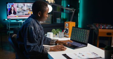 Merkez ofisteki serbest çalışan borsa yatırımcısı dizüstü bilgisayardaki borsa kârını kontrol ediyor. Apartmanda serbest çalışan Afrikalı Amerikalı piyasa hisseleri, not defteri, kamera B