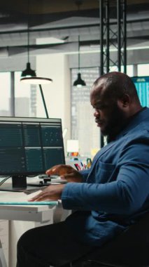 Modern çalışma alanındaki dikey Video Siyah Erkek Yöneticisi bir bilgisayar monitöründe öngörü ve iş stratejisini analiz ederek, üretkenlik için rapor yazmayı ve planlamayı güvence altına alır. Kamera A.