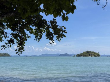 Tayland 'da küçük bir adanın kıyılarında yüzen ahşap bir tekne kristal berrak turkuaz sularda bir yelkenli teknesinin bulunduğu manzaralı bir deniz manzarası. Bereketli tropikal manzara ve parlak mavi gökyüzü.