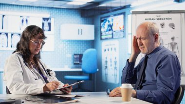 Kadın doktor, kontrol sırasında migreni tedavi etmek için uzman tavsiyesi veriyor. Doktor, radyoloji raporları ve reçeteli ağrı kesicilerle modern bir dolapta yaşlı bir hastaya danışıyor. Kamera A.