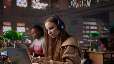 Kampüs kütüphanesinde kulaklıkla konferansa katılan genç bir öğrenci, notlar alıyor ve online bir toplantıda politika hakkında bilgi ediniyor. Ders sırasında odaklanmış bir öğrenci. Kamera B.