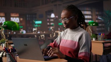 Üniversite kütüphanesindeki veritabanından veri toplayarak, daha fazla akademik bilgi toplamak için araştırma araçları kullanarak edebiyat ödevine odaklanan genç zenci öğrenci. Kamera B.