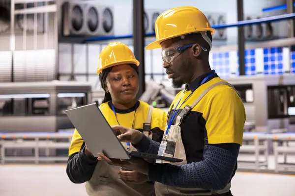 Endüstri 4.0 uzmanları akıllı üretim fabrikasındaki robot kol üretim hattını denetleyerek kusursuz otomasyonu sağlıyorlar. Siyah mühendisler otomatikleştirilmiş fabrikada dizüstü bilgisayarla dijital optimizasyon yapıyorlar..