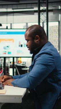 Dikey Video Afrikalı Amerikalı analist bir ders kitabına büyüme stratejisi notları yazıyor, iş ofisinde anlayış ve finansal verilerle çalışıyor. Operasyonel başarı ve verimliliği güvence altına alan danışman