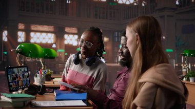 Çeşitli öğrencilerden oluşan bir grup, kampüs kütüphanesindeki uzak sınıf seminerine katılmak üzere çevrimiçi kurslara katılıyor. Gençler akademik hocadan notlar alıyorlar. Kamera B.