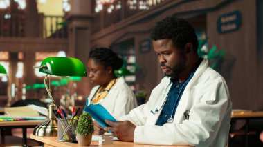 Tıp öğrencisi bir kitap okuyor ve kütüphane veritabanından tıbbi araştırmalara odaklanmış notları inceliyor. Genç adam, sağlık kariyerine ve akademik hedeflere olan bağlılığını yansıtıyor. Kamera B.