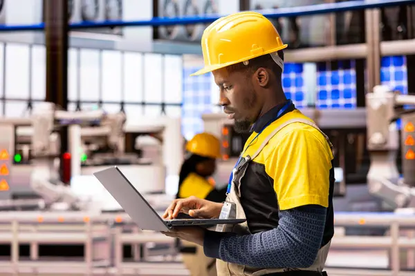 Dizüstü bilgisayarı olan erkek mühendis akıllı üretim makinelerini denetliyor, otomatik fabrika operasyonlarını güvence altına alıyor. Afrikalı Amerikalı teknisyen endüstri 4.0 sisteminin optimizasyonu için dijital arayüz kullanıyor.