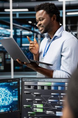 Happy Veri Merkezi teknisyeni, yapay zeka sinirsel ağlarını görüntülemek için laptop aygıtı kullanıyor. Sunucu odasındaki neşeli adam defter ekranına bakıyor, yapay zeka sistemlerini denetliyor.