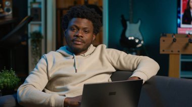 Neşeli Afrikalı Amerikalı adam şık bir oturma odasında dizüstü bilgisayarıyla telgraf çekiyor. Modern esnek çalışma alanında konfor ve üretkenliği dengeleyen uzaktan kumandalı bir işçi. Rahat bir çatı katı stüdyosu. Kamera A.