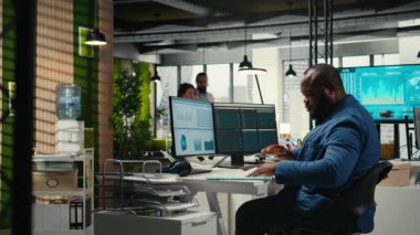 Modern çalışma alanındaki siyah erkek yönetici bir bilgisayar monitöründe öngörü ve iş stratejisini analiz ederek, üretkenlik için rapor yazmayı ve planlamayı güvence altına alır. Kamera A.