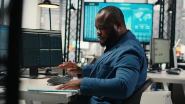 Modern çalışma alanındaki siyah erkek yönetici bir bilgisayar monitöründe öngörü ve iş stratejisini analiz ederek, üretkenlik için rapor yazmayı ve planlamayı güvence altına alır. Kamera B.