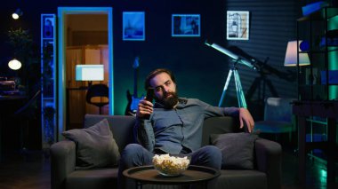 Televizyon ekranındaki kanalları değiştirmek için uzaktan kumanda kullanan bir adam ilginç olmayan içerikten bıkmış hisseder. TV kanallarında gezinen sıkkın insan izlenebilir bir şey bulamayınca üzülüyor, B kamerası.