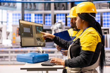 Siyah mühendislik ekibi montaj hattındaki robotik güneş paneli üretimini kontrol etmek için masaüstü bilgisayarı kullanıyor. Sanayi 4.0 üretim sistemleri ve endüstriyel otomasyon hız ve kesinliği artırıyor.