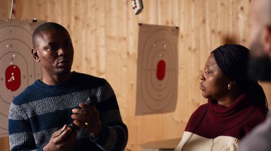 Atış poligonu güvenlik görevlisi müşterilere nasıl silah doldurulacağını gösteriyor. Kurallara uymalarını sağlıyor. Atış poligonu çalışanı düzgün silah kullanmayı ve kullanmayı öğretiyor, kamera B