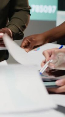 İş hedeflerini modern ofis alanındaki beklentilerle aynı hizaya getirmek için geç saatlerde düzenlenen bir şirket brifinginde, analizleri, projeksiyonları ve veri içgörülerini gözden geçiren dikey video çok ırklı bir ekip. Kamera.