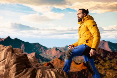 Yürüyüşçü dramatik dağ zirveleri ve bulutların panoramik manzarasına bakar, maceranın heyecanını ve azmin ödüllerini hisseder. Yolculuk gücü ve kişisel gelişimi temsil eder..