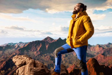 Yürüyüşçü, vahşi doğada yürümenin zorluğunu kucaklıyor, panoramik dağ manzarasının tadını çıkararak, gücün ve açık hava macerasının ödüllerini anımsatıyor. Macera Avcısı.