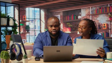 Aylık rapor numaralarını dizüstü bilgisayar ve kağıtlarla değerlendiren, rakamları analiz eden ve son teslim tarihinden önce bakiyeyi hesaplayan genç bir çift. Evden çalışan Afrikalı Amerikalılar. Kamera A.