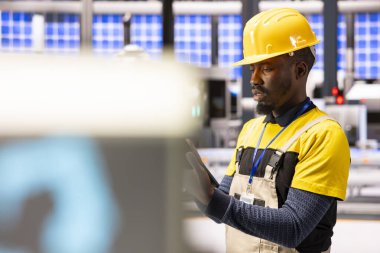 Otomatik fabrikadaki siyah erkek mühendis Endüstri 4.0 sistemini izliyor. Afrikalı Amerikalı teknisyen tablet üzerinde çalışıyor, endüstriyel otomasyonu geliştiriyor, akıllı üretim süreçlerini güvence altına alıyor.