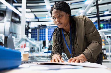 Siyah kadın güneş paneli makinelerini kontrol ediyor ve dosyalardaki kontrol listesini gözden geçiriyor, yenilenebilir enerji üretim ve montaj hattında hayati bir rol oynuyor. Verimli güneş teknolojisi altyapısı.