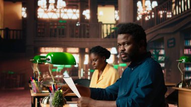 Kampüs kütüphanesinde sınavlara hazırlanan kararlı öğrenci, tam bilgi için kitaplar, notlar ve araştırma belgeleri gibi eğitim kaynaklarını kullanıyor. Üretkenlik için sessiz bir ortam. Kamera A.