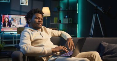 Genç bir yetişkin, dairesinden dizüstü bilgisayarla çalışıyor. Siyah serbest çalışan, rahat bir kanepeden internette gezinirken verimlilik ve rahatlığı harmanlayan görevlere odaklanıyor. Kamera B.