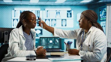 Klinikteki rutin kontroller sırasında sıcaklığı ölçen siyah sıhhiye grip belirtilerini kontrol etmek için dijital termometre kullanıyor. Sağlık uzmanı bir hastaya teşhis koymayı garanti ediyor. Kamera B.