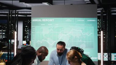 İş arkadaşları, strateji odasındaki belgeleri inceleyerek, raporları değiş tokuş ederek ve şirket yönetimine ilişkin tahminleri sunarak sunuma hazırlanıyorlar. Kamera A.