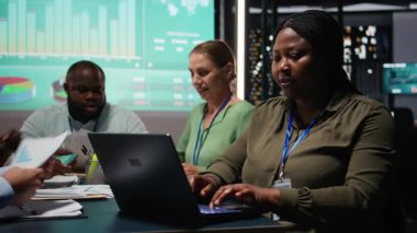 Afrikalı Amerikalı analist ve meslektaşları, küresel iş gelişimini kolaylaştırmak için gece yapılan bir toplantıda yıllık veri raporlarını inceliyor. Çalışma takımı. Kamera A.