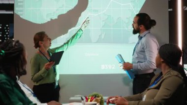 Gece yarısı şirket toplantısı sırasında yeni bir proje gündemi için veri analizi ve içgörülerini paylaşan bir kuruluştan çeşitli personel. İşçiler kaynak tahsisi konusunu tartışıyor. Kamera B.