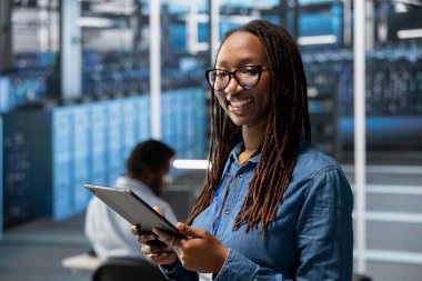 Gülümseyen sunucu merkezi bilişim uzmanı vites sorunlarını çözüyor. Dar geçitleri tanımlamak için ekipman performansını izlemek için tablet kullanan radyan veri merkezi sistem yöneticisinin portresi.