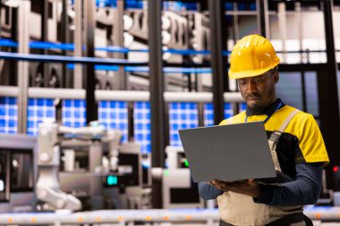 Siyah mühendis endüstriyel santraldeki 4.0 endüstriyel sistemi izliyor. Robot kollar ve taşıma bantlarıyla çevrelenmiş halde, akıllı imalata odaklanarak otomatikleştirilmiş fabrika optimizasyonunu sağlamak için dizüstü bilgisayar kullanıyor.