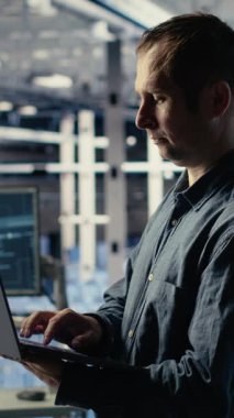 Veri merkezindeki dikey video mühendisi, rutin sistem incelemesi sırasında dizüstü bilgisayarı kullanarak Al 'ın gösterge panelini kontrol ediyor. IT uzmanı güvenilir performansı doğrulamak için yapay zeka araçlarını not defterinde test ediyor, kamera