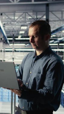 Veri merkezindeki dikey video mühendisi, rutin sistem incelemesi sırasında dizüstü bilgisayarı kullanarak Al 'ın gösterge panelini kontrol ediyor. IT uzmanı güvenilir performansı doğrulamak için yapay zeka araçlarını not defterinde test ediyor, kamera