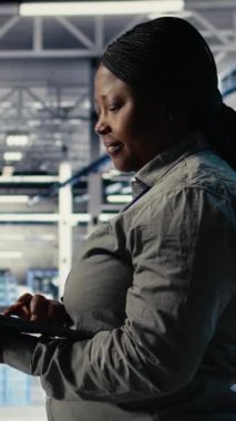 Dikey video bilişim uzmanı tesiste yürürken dizüstü bilgisayarla sunucu çiftlik etkinliğini izliyor. Veri merkezindeki Afrikalı Amerikalı kadın sistemlerin doğru çalıştığından emin olmak için test sonuçlarını gözden geçirdi