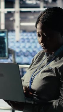 Dikey video veri merkezi yönetimi, teçhizat sistemleri incelemesi sırasında dizüstü bilgisayarla Al 'ın gösterge panelini kontrol ediyor. Sunucu merkezindeki Afrikalı Amerikalı kadın güvenilir olup olmadığını kontrol etmek için yapay zeka araçlarını bilgisayarda test ediyor.