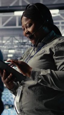 Dikey video sunucusu merkezi teknik destek uzmanı şirket müşterilerine güvenli veri yedekleri kurma konusunda rehberlik ediyor. Veri merkezindeki Afrikalı Amerikalı kadın kullanıcıların sunuculardaki sistem hatalarını düzeltmelerine yardım ediyor