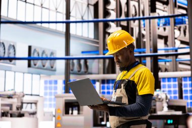 Profesyonel erkek mühendis yazılım sistemlerini dizüstü bilgisayarda düzelterek endüstriyel santraldeki otomatikleştirilmiş üretim hatlarını optimize ediyor. Robot kollar, akıllı fabrikadaki teknoloji ve yeniliğin birleşimini vurguluyor.