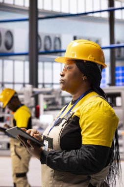 Siyahi kadın teknisyen montaj hattını denetliyor ve en iyi performansı sağlamak için tablet kullanıyor. Kadın mühendis otomatik fabrika sistemini izliyor, işlevsellik sektörünü denetliyor 4.0 yazılım.