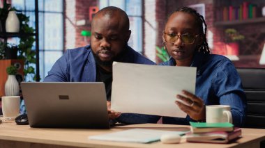 Aylık rapor numaralarını dizüstü bilgisayar ve kağıtlarla değerlendiren, rakamları analiz eden ve son teslim tarihinden önce bakiyeyi hesaplayan genç bir çift. Evden çalışan Afrikalı Amerikalılar. Kamera B.