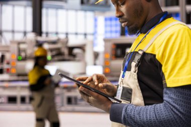 Siyah erkek mühendis akıllı üretim tesisinde dijital otomasyon sistemlerini izliyor ve tablet kullanarak endüstriyel bitki ekipmanlarını optimize ediyor. Otomatik fabrika uzmanı üretim sürecini denetliyor.